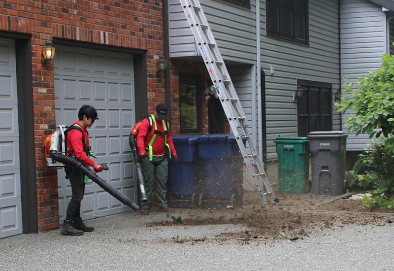 Two gutter specialists from a gutter company serving King and Snohomish counties in Washington blow leaves next to a brick three car garage.